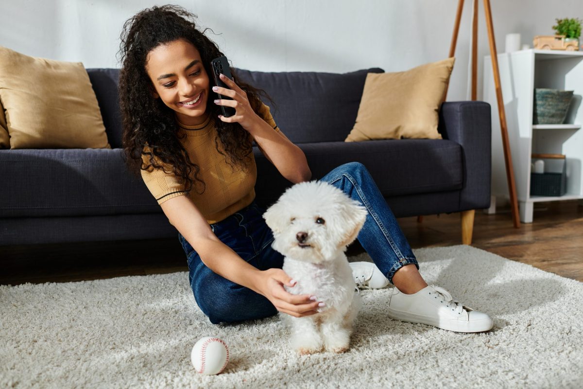 Woman with curly hair sitting on floor, petting white Bichon Frise dog lovingly.
